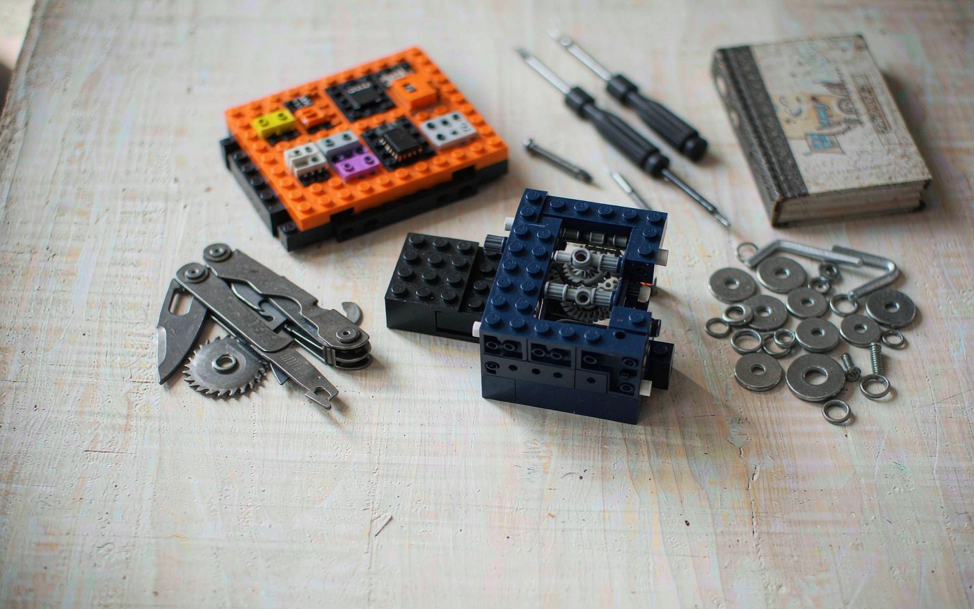 Flat lay of a metal construction set, snap-together electronics board, brick gear mechanism, multitool and precision screwdrivers on a pale oak table